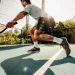 A person in athletic clothing and pickleball shoes is playing pickleball outdoors on a sunny day. They are holding a paddle and are in a low, athletic stance on a blue court with a white dividing line, with trees and a fence in the background.